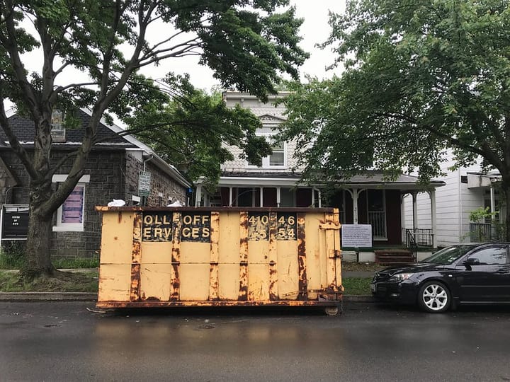 A large yellow but rusting construction dumpster on the street in front of a 2 storey house with a porch.