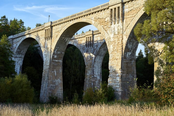 Two stone railway bridges in a sparse forest with tall, wild grass in the foreground.
